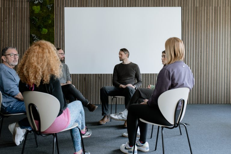 Eine Gruppe von Menschen sitzt im Kreis auf Stühlen in einem modernen Raum und nimmt an einer moderierten Gruppendiskussion oder Therapiesitzung teil.