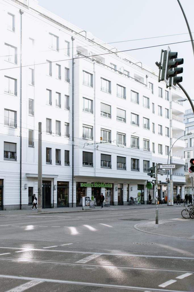 Eine helle Außenaufnahme eines modernen, mehrstöckigen weißen Gebäudes an einer städtischen Straßenecke. Im Erdgeschoss befindet sich ein „Denns BioMarkt“. Im Vordergrund sind Straßenbahnschienen und eine Verkehrsampel zu sehen, was auf eine zentrale und gut erreichbare städtische Lage hindeutet.