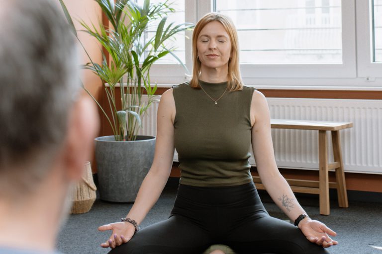 Eine blonde Frau sitzt im Meditationssitz auf einer Yogamatte, die Augen geschlossen und die Handflächen nach oben gerichtet. Sie wird aus der Perspektive eines anderen Teilnehmers im Vordergrund (unscharf) gezeigt. Der Raum ist durch ein großes Fenster lichtdurchflutet und mit warmen terracottafarbenen Wänden sowie einer großen Zimmerpflanze gestaltet.