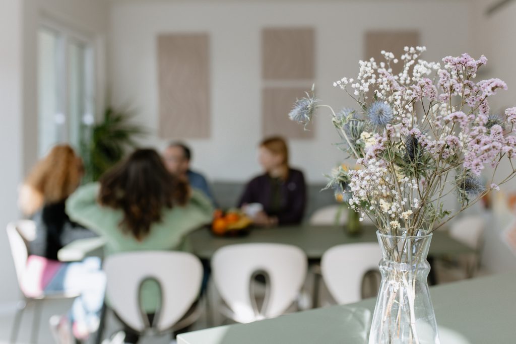 Ein heller, luftiger Raum mit einer gläsernen Vase voller getrockneter Wildblumen im Vordergrund. Im unscharfen Hintergrund sitzt eine Gruppe von Menschen an einem grünen Tisch und unterhält sich. Der Raum ist mit modernen weißen Stühlen ausgestattet und wirkt sauber und therapeutisch einladend.