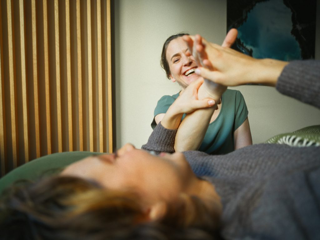 Eine Therapeutin mit zusammengebundenen Haaren lächelt eine Patientin (unscharf im Vordergrund) herzlich an. Die Therapeutin hält und führt sanft den Arm der Patientin während einer Sitzung. Im Hintergrund ist eine minimalistische Wand mit hellen vertikalen Holzlamellen zu sehen.