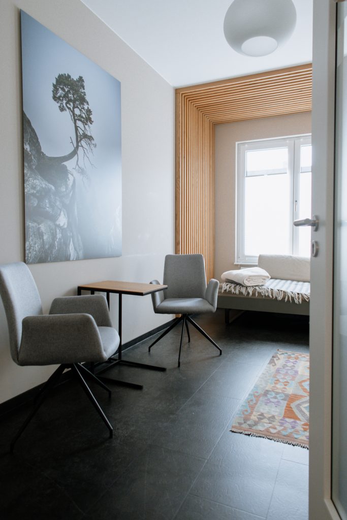 A view looking into a bright consultation room through a partially open door. The room features two grey upholstered swivel chairs facing a small wooden side table. On the left wall hangs a large canvas print of a lone tree on a rocky, misty cliff. In the background, a therapy bed with blankets sits by a window framed with vertical wooden slats.